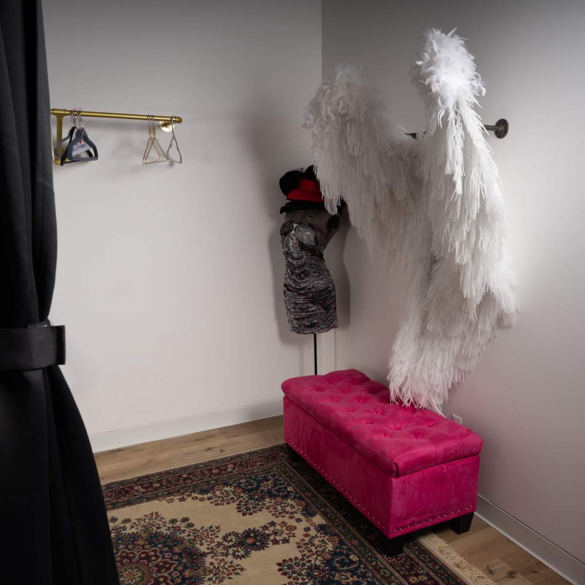 A small dressing room features a pink tufted bench, a mannequin displaying a dress, and large white feather wings hanging on the wall.
