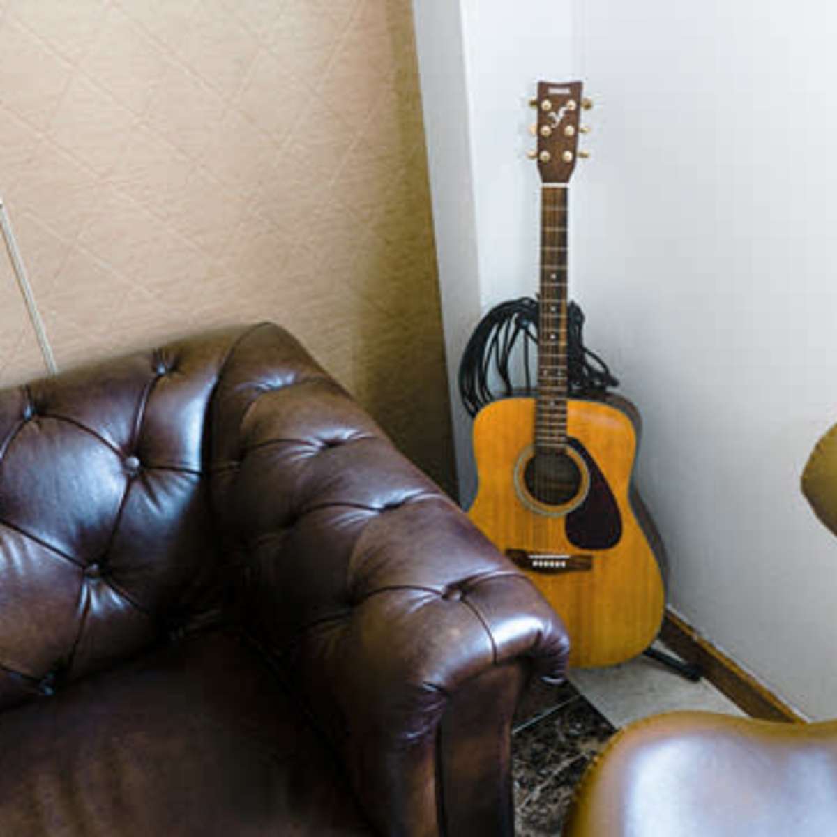 A wooden acoustic guitar stands in the corner next to a leather couch and an armchair in a well-lit room.
