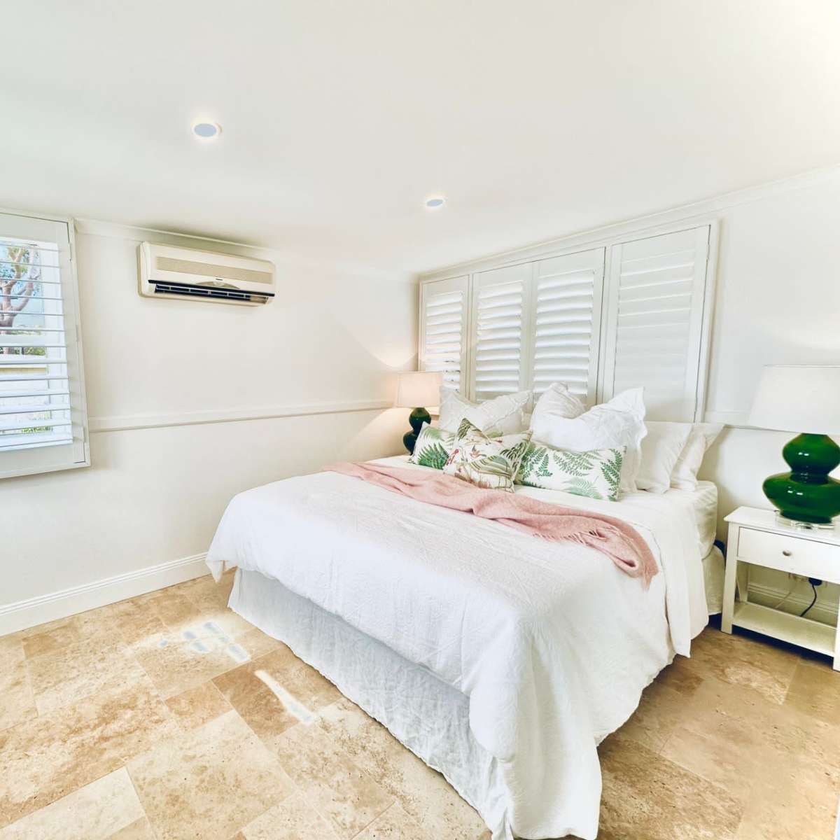 A bright bedroom featuring a double bed with decorative pillows, a small round dining table, and windows with plantation shutters.