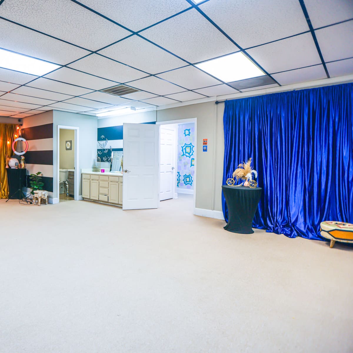 A spacious room with beige carpet, featuring colorful curtains in gold and blue, a lounge chair, and various decorative items, along with doors leading to other areas.
