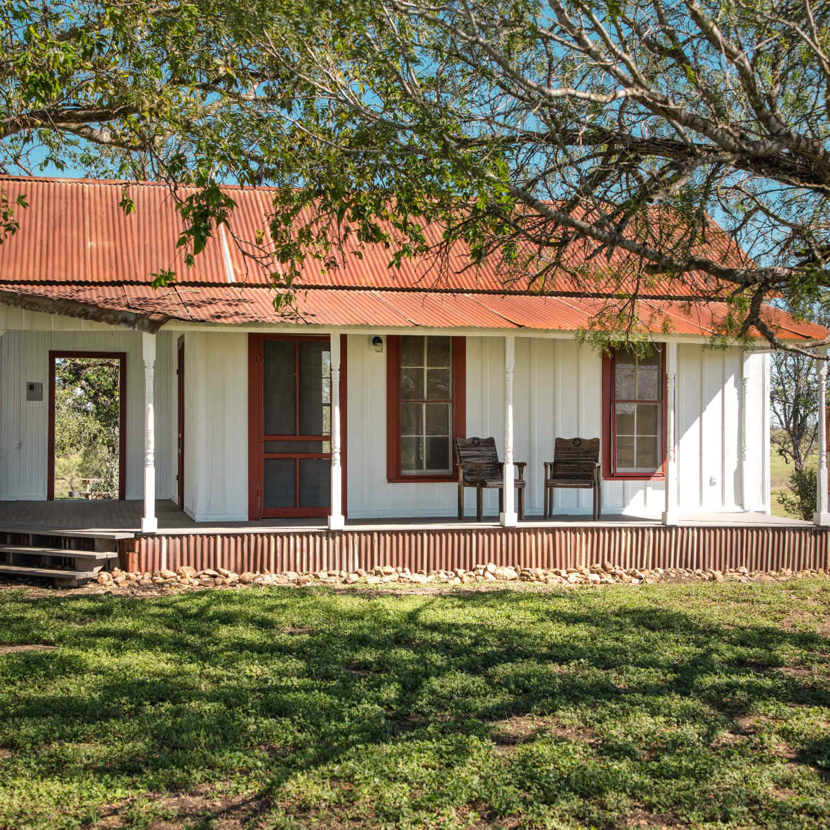 A white, wooden house with a red tin roof and two rocking chairs on the porch stands amid a grassy area, accompanied by a nearby round metal tank.