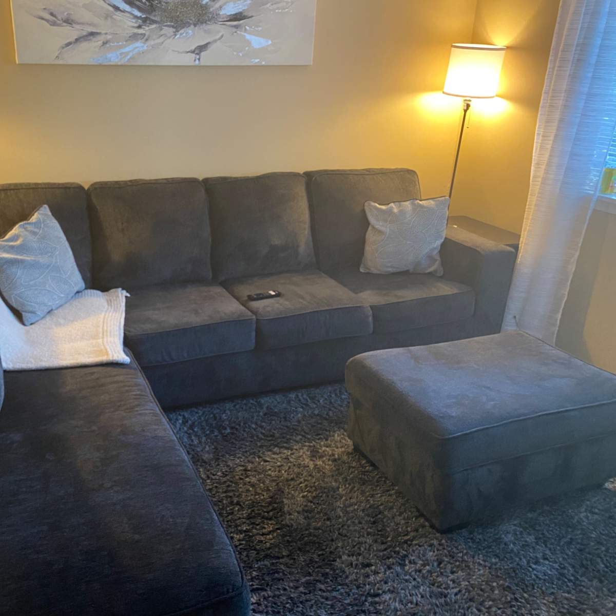 A sectional sofa with pillows and a throw blanket is positioned in a well-lit living room with two lamps and a matching ottoman.