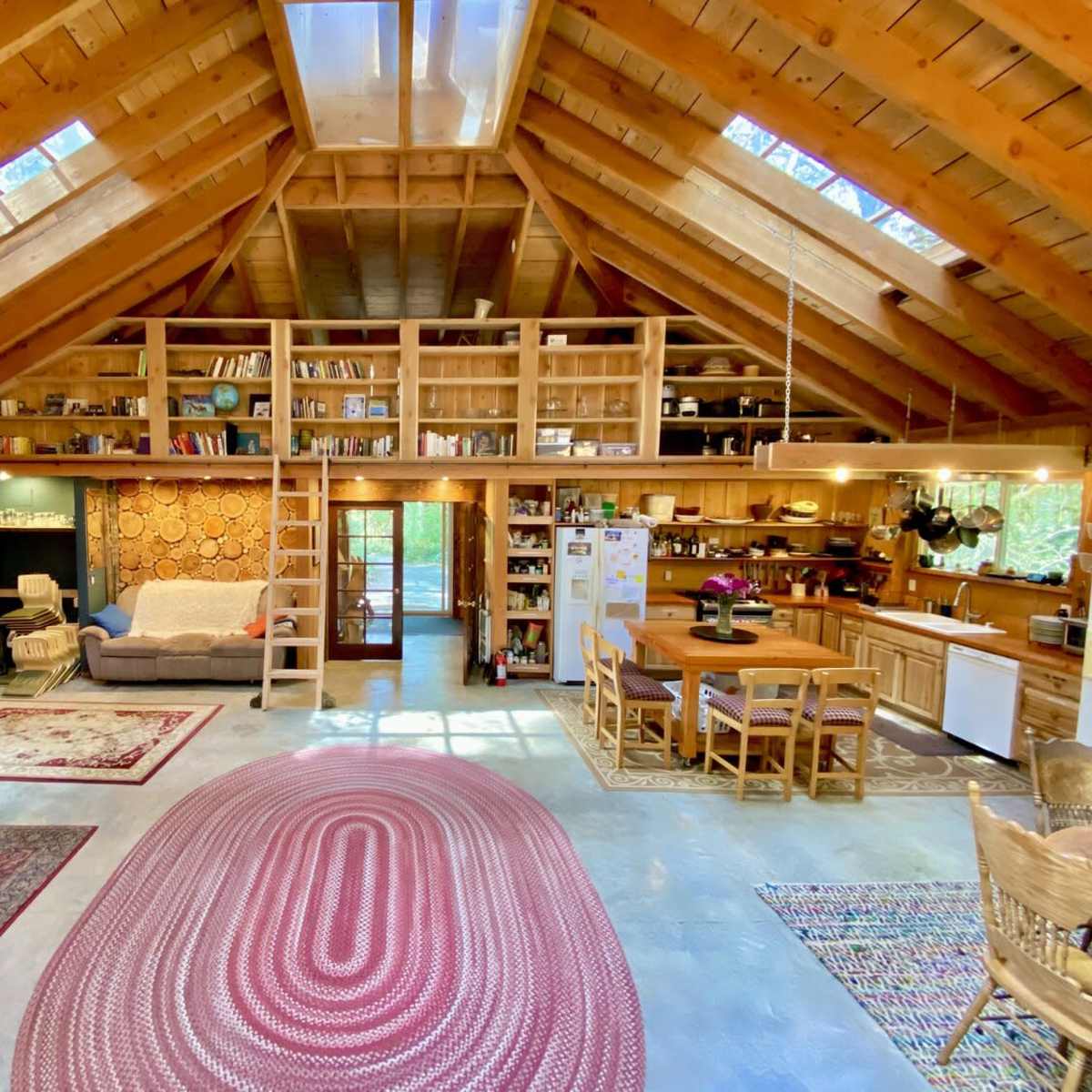 A spacious interior of a rustic cabin with wooden beams, bookshelves, a dining area, and a cozy seating area.