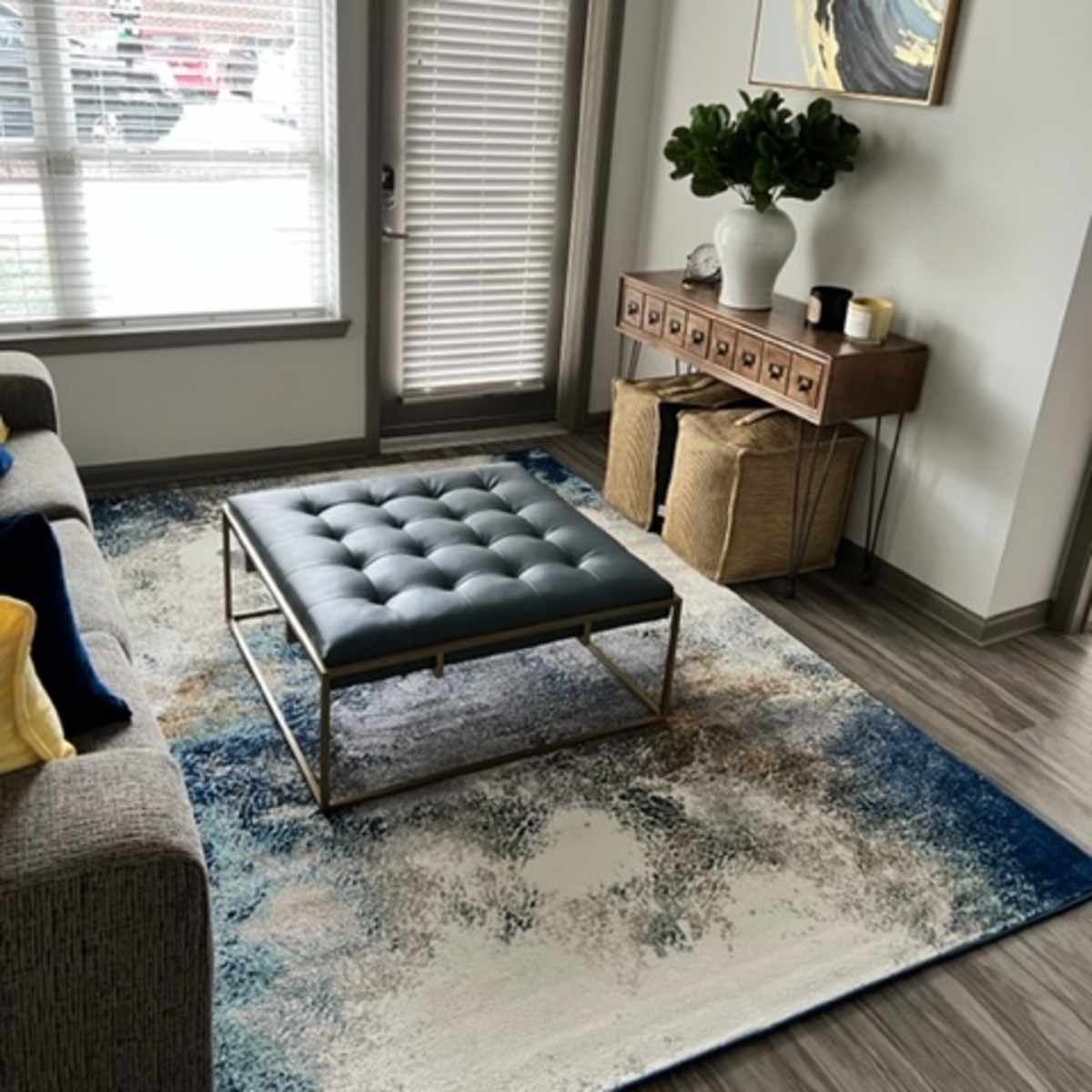 A modern living room with a gray sofa, a blue tufted ottoman on a decorative area rug, and a wooden side table with a plant and a decorative item.
