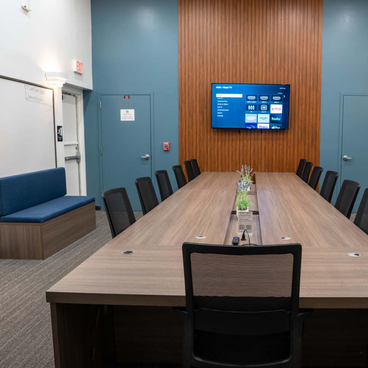 A long conference table with chairs surrounds a screen mounted on a wooden wall in a modern meeting room.