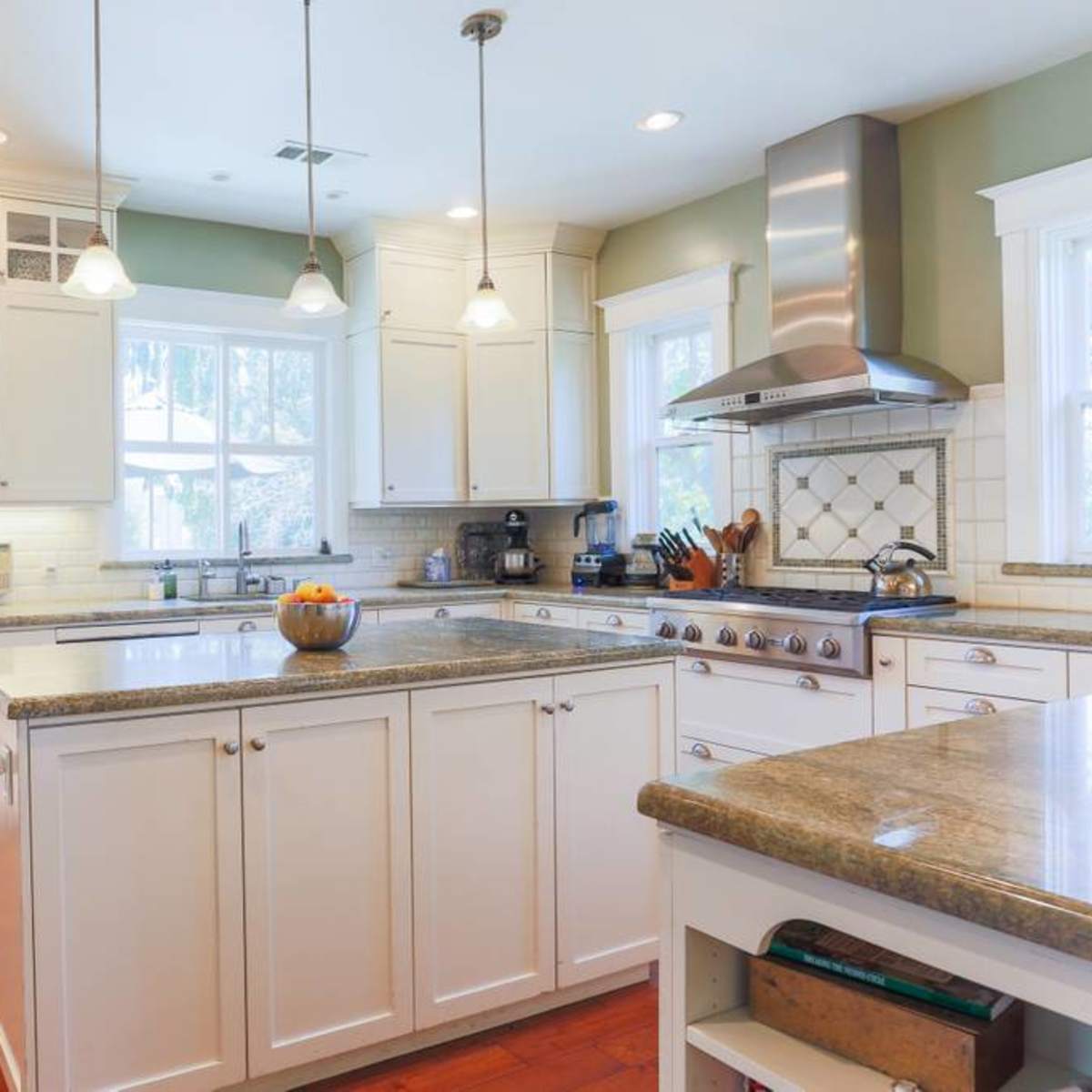 A bright kitchen features white cabinetry, a gray stone island with a bowl of fruit, and wooden flooring.