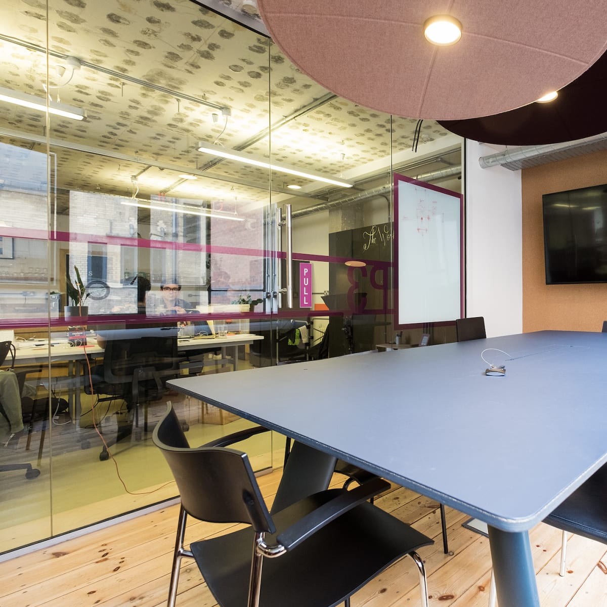 A modern conference room features a large table surrounded by chairs, a wall-mounted screen, and glass partitions with pink accents.