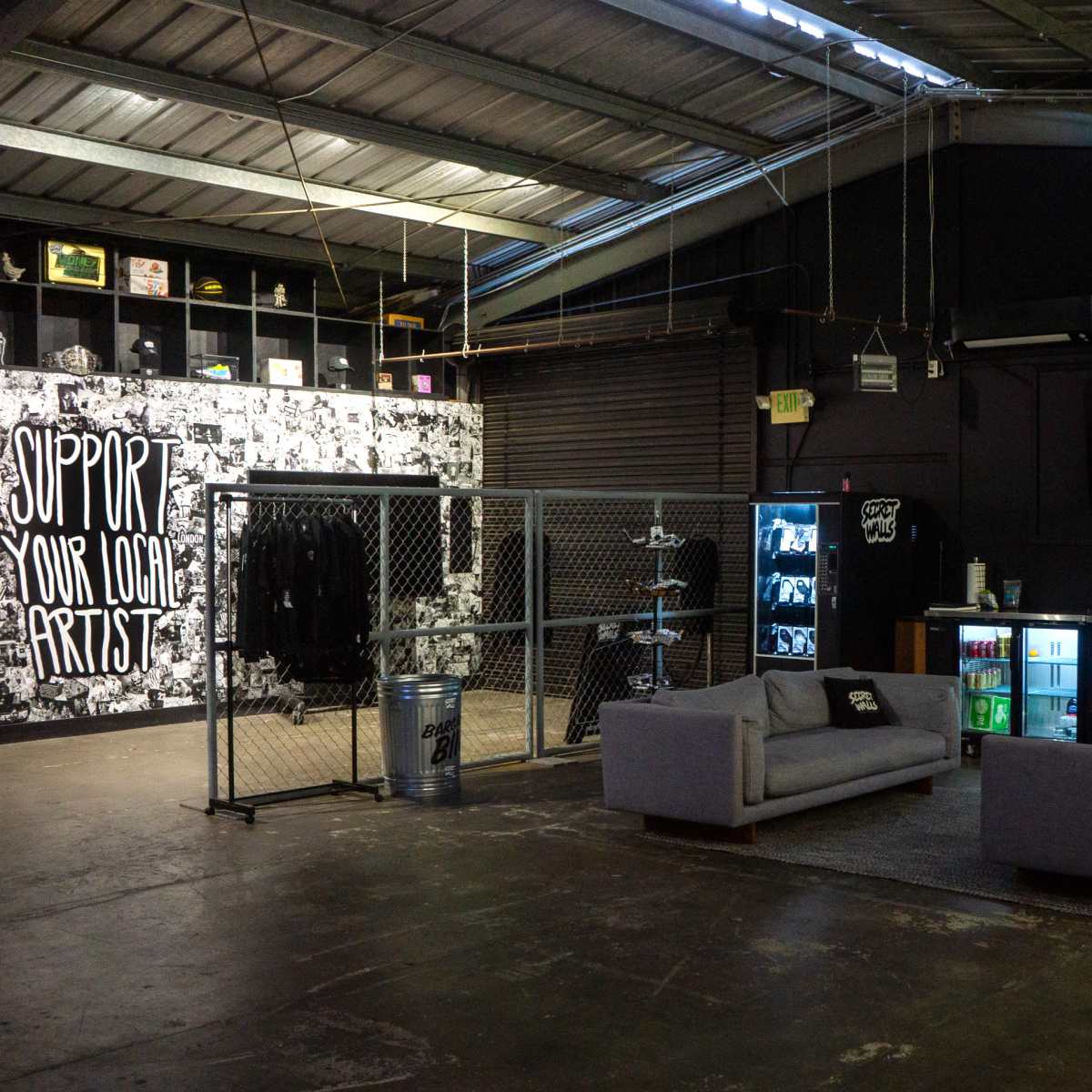An industrial-style lounge area with a chain-link fence, two gray sofas, and a wall featuring artwork that encourages support for local artists.