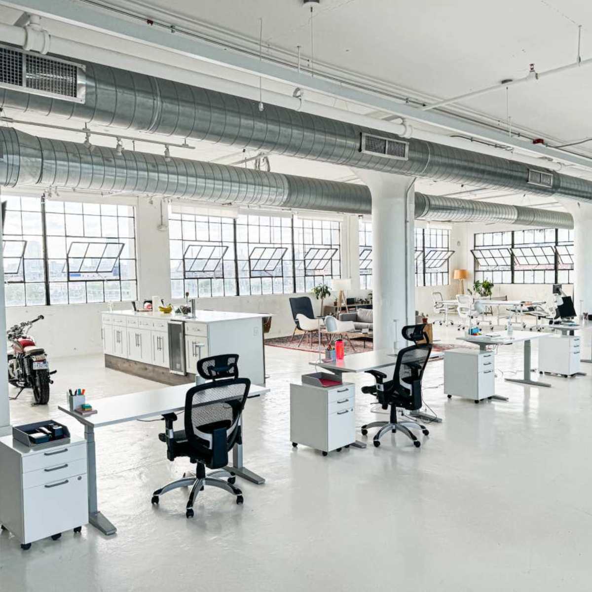 A spacious modern office with several desks, ergonomic chairs, and large windows, along with a kitchenette in the background.