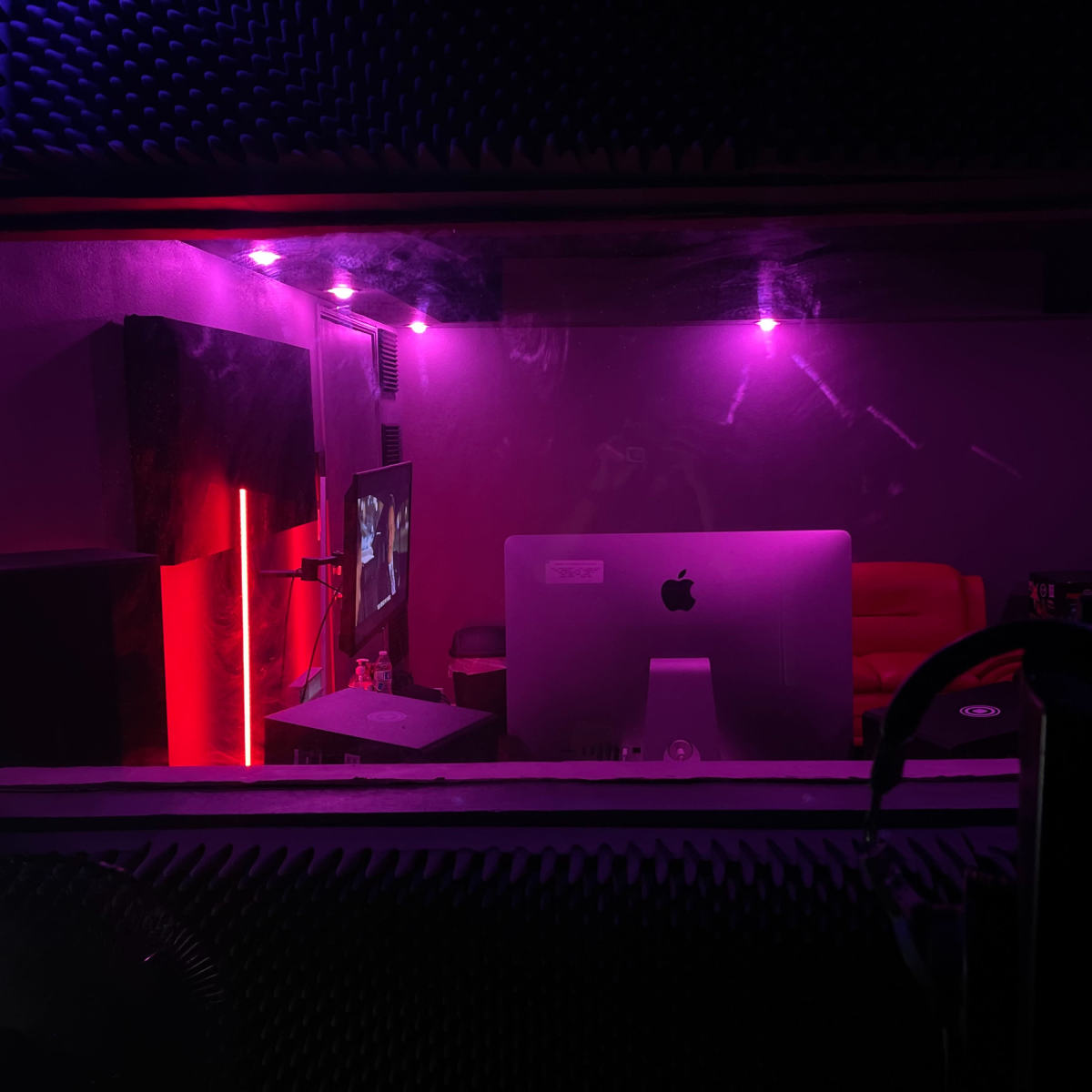 A dimly lit recording studio with purple lighting, featuring an iMac, a couch, and soundproofing elements on the walls.