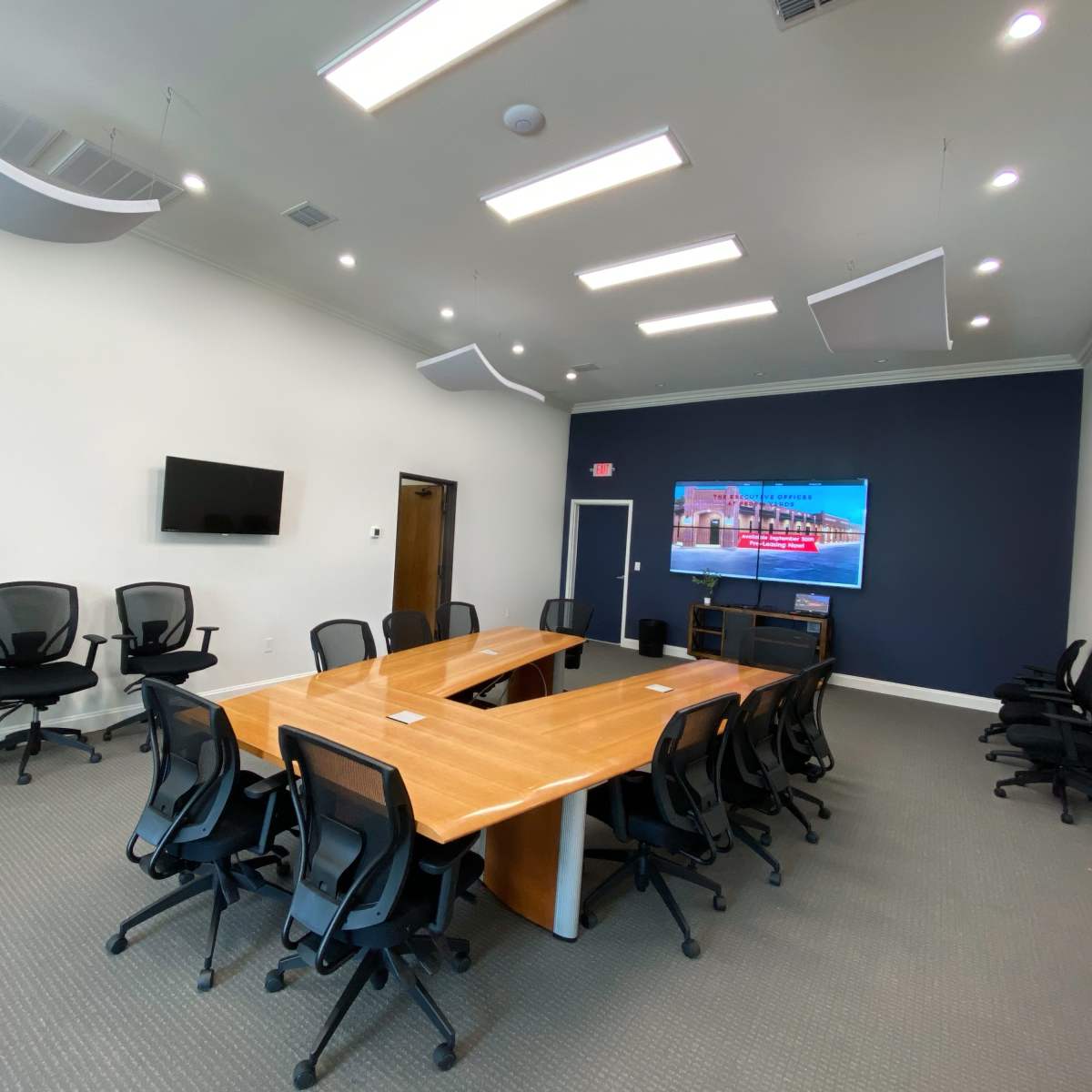 A modern conference room features a large wooden table surrounded by black ergonomic chairs, with a wall-mounted television and a minimalist design.