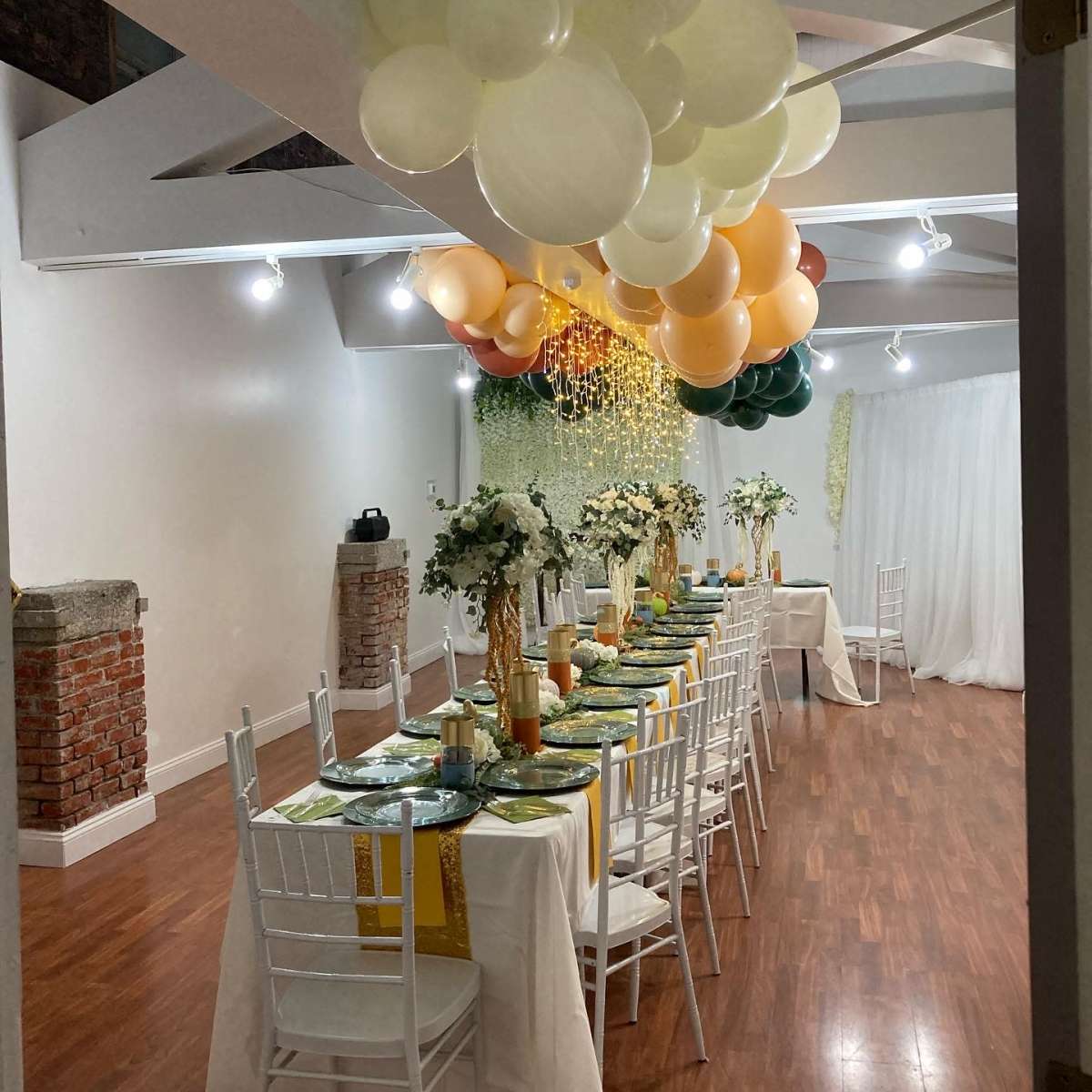 A long dining table is set with green and gold decorations, surrounded by white chairs, beneath a ceiling adorned with a cluster of balloons.