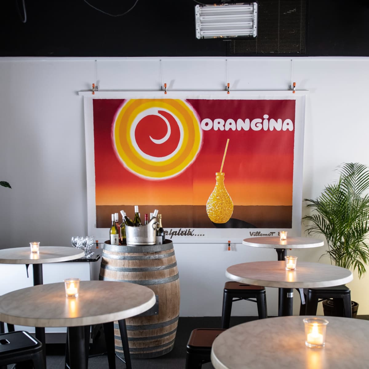 A brightly colored Orangina advertisement hanging on the wall of a casual dining area featuring round tables and a wooden barrel.