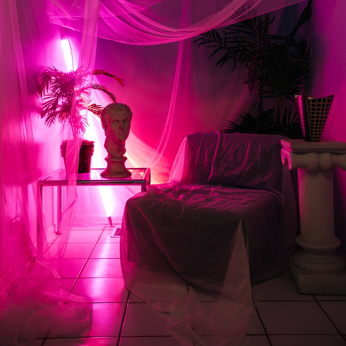 A dimly lit room features a transparent drapery setup, a bust on a pedestal, a small table with a plant, and a chair covered in fabric.