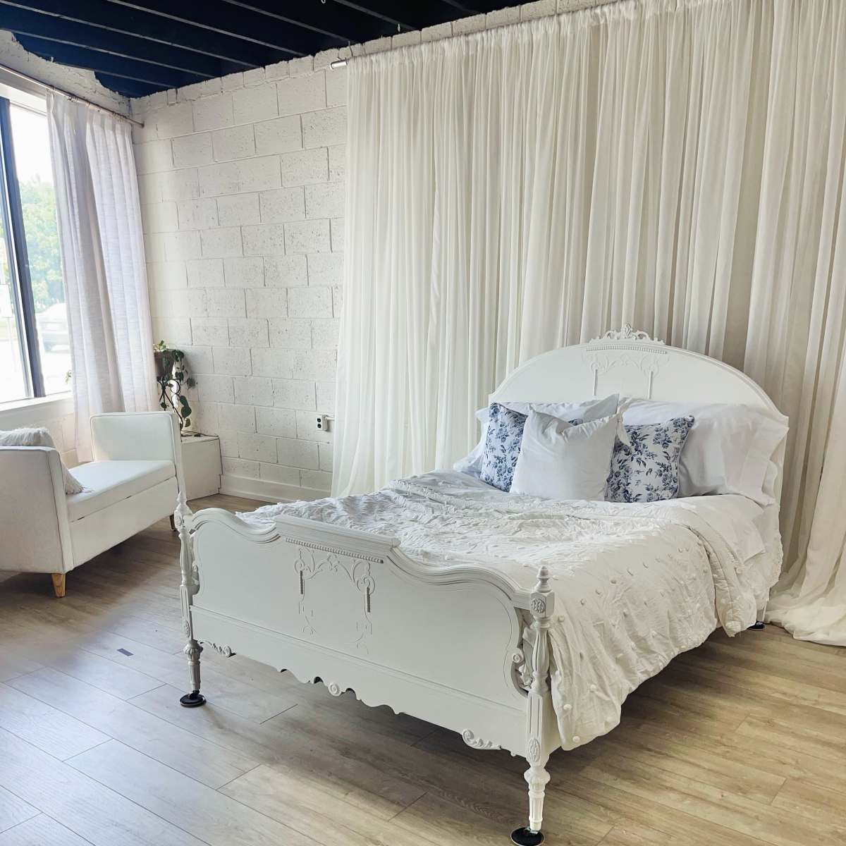 A white, ornate bed with a light-colored bedding set positioned in a room with large windows and sheer curtains.