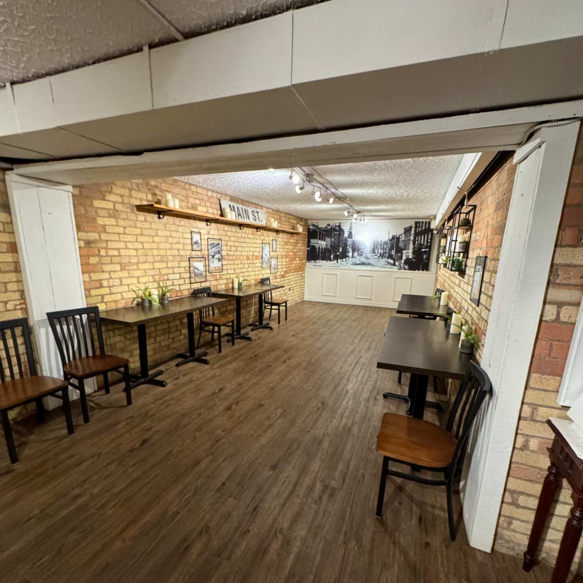 An indoor cafe or restaurant with wooden tables and chairs arranged along a brick wall, featuring framed pictures and a decorative backdrop.