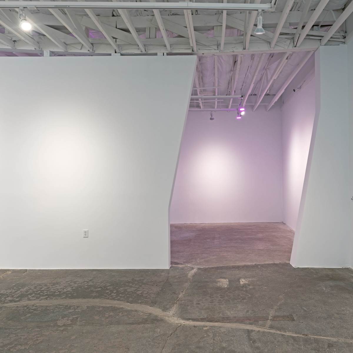 A minimalist interior space with white walls, a concrete floor, and an angled partition leading to another room.