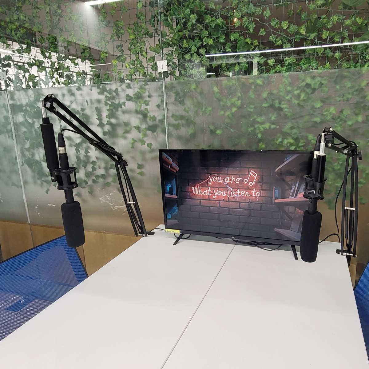 A television displaying a neon sign surrounded by two microphone stands is positioned on a table in a room with greenery on the walls.