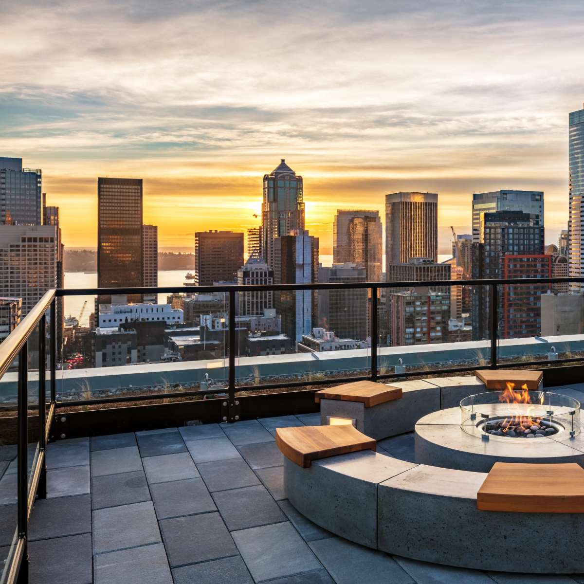 A city skyline at sunset is viewed from a rooftop terrace featuring a circular fire pit surrounded by wooden seating.