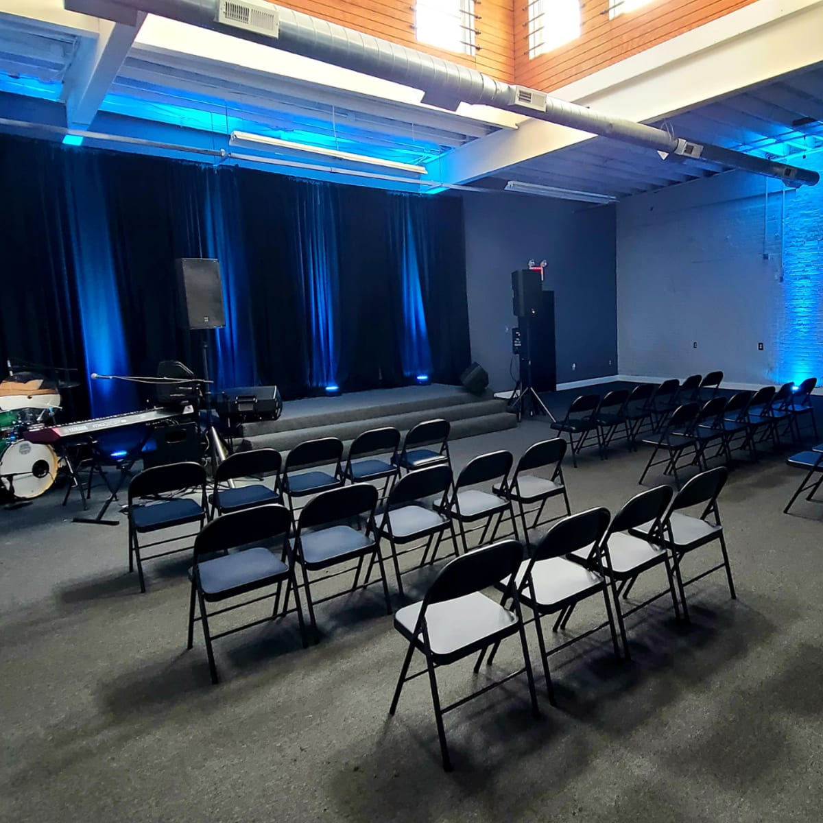 A performance space with black chairs arranged in rows facing a small stage, flanked by blue-lit curtains and a drum set in the corner.