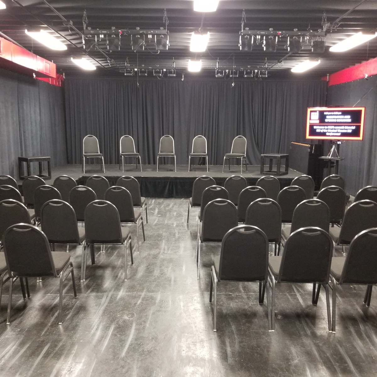 A minimalist stage is set with six chairs arranged on a platform, facing an audience area filled with black chairs, against a backdrop of black curtains and overhead lights.