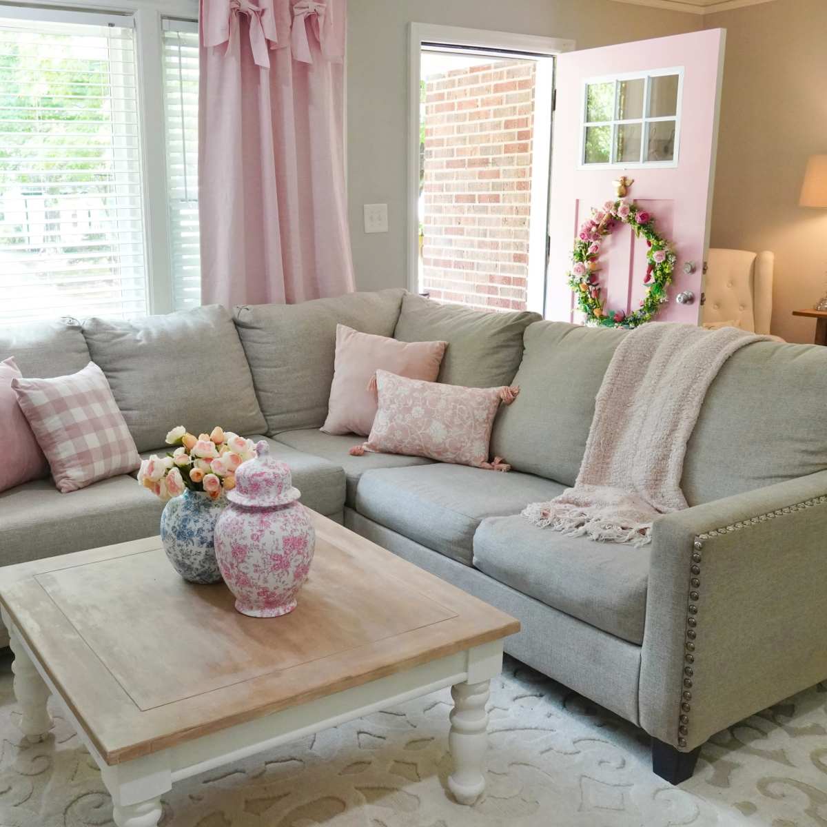 A cozy living room featuring a gray sectional sofa adorned with pink decorative pillows, a light coffee table, and a vase of flowers on top.