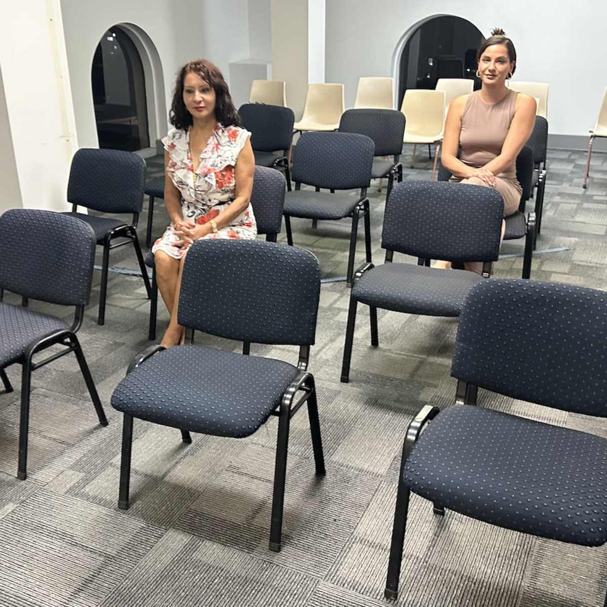 Two women sit in an empty room with rows of chairs arranged around them.