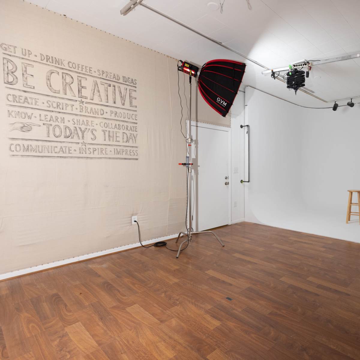 A photography studio features a white backdrop, wooden flooring, and a stool in the center, with lighting equipment set up along one side.