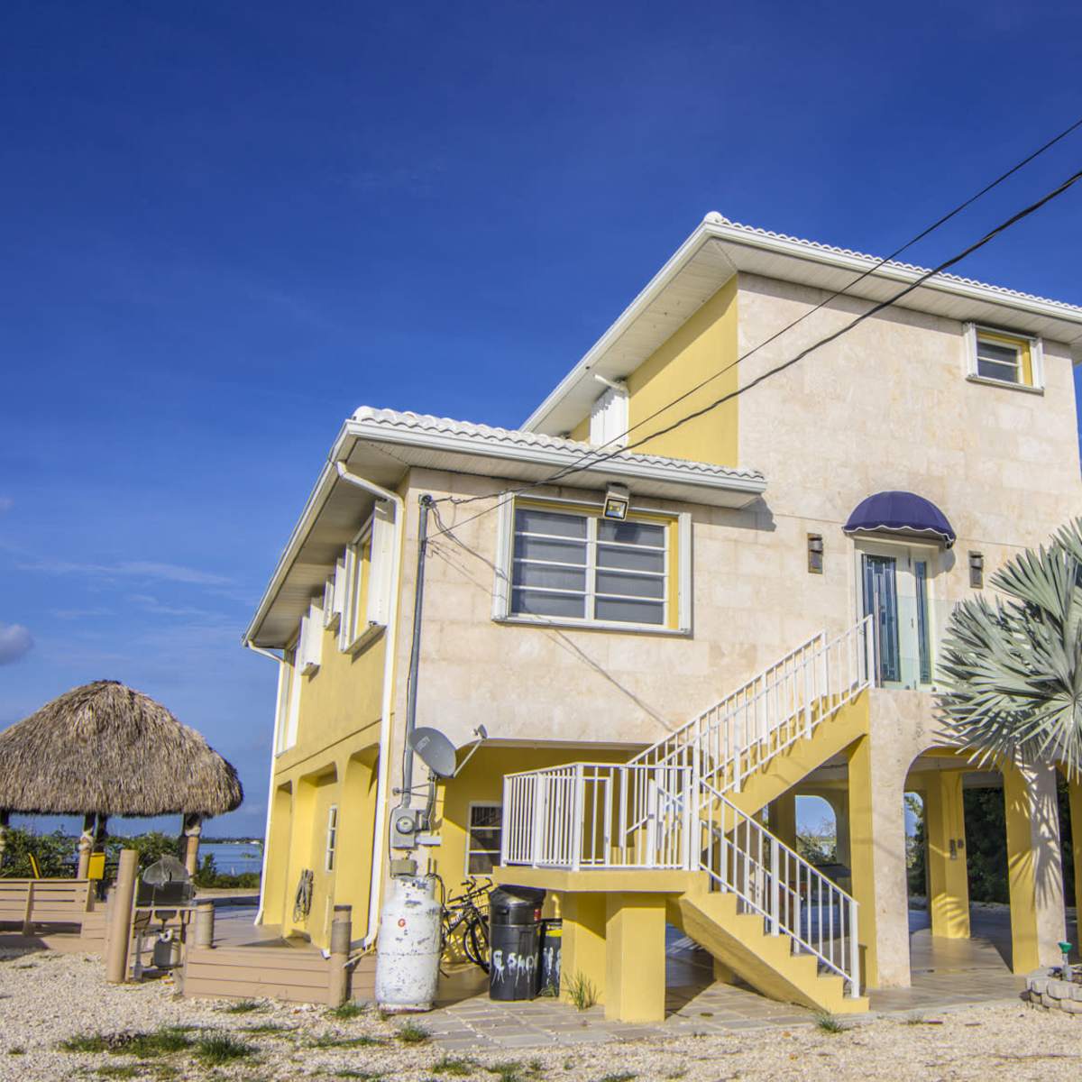 A two-story yellow house with a porch and a thatched-roof gazebo nearby, set against a clear blue sky.