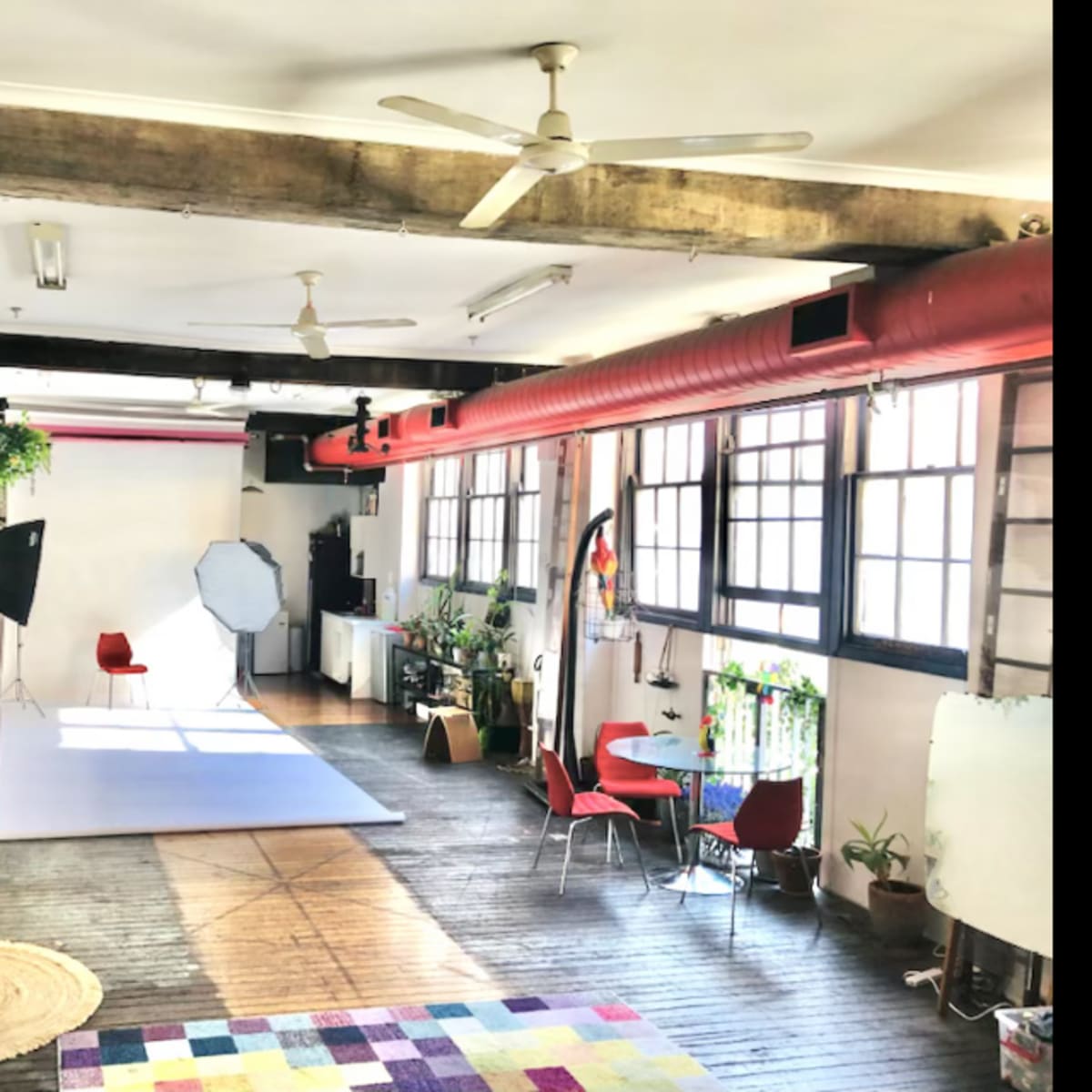 A spacious, well-lit studio featuring wooden floors, large windows, plants, and various seating areas, including a red chair and a rug.