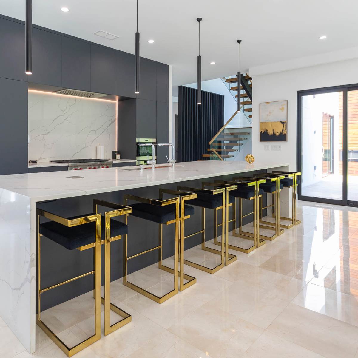 A modern kitchen with a large marble island, black cabinetry, and gold-accented bar stools.