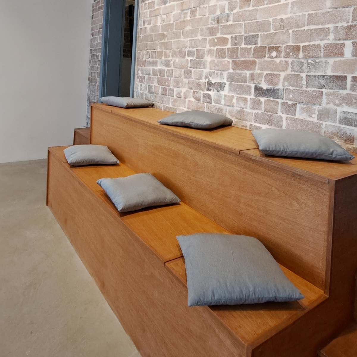 A minimalist seating area with wooden steps and gray cushions against a textured brick wall.