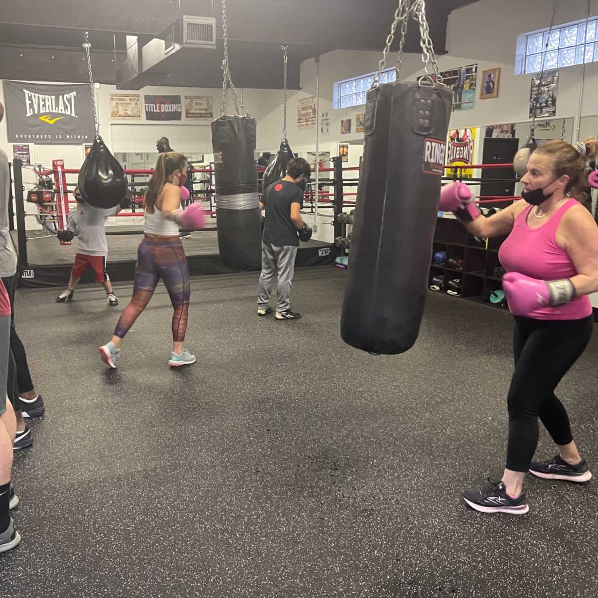 A group of people exercises in a boxing gym, using punching bags and wearing gloves and masks.