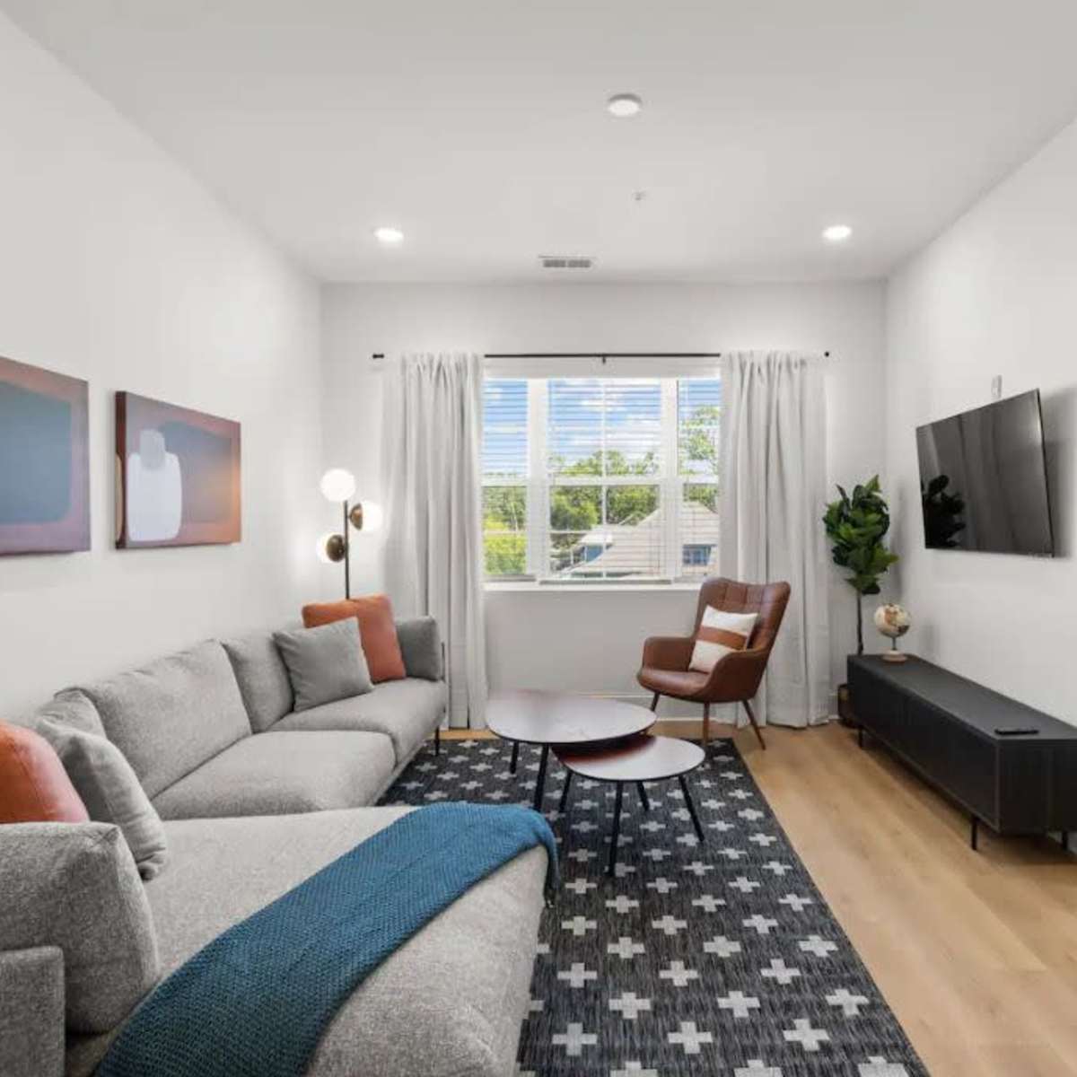 A modern living room features a gray sectional sofa, a round coffee table, and artworks on the walls, with a window offering natural light and a view outside.