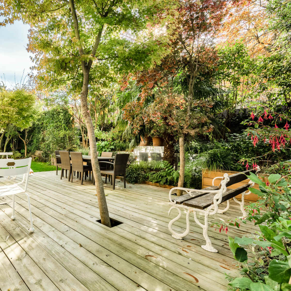 A wooden deck surrounded by greenery, with several seating areas and a dining table under trees.