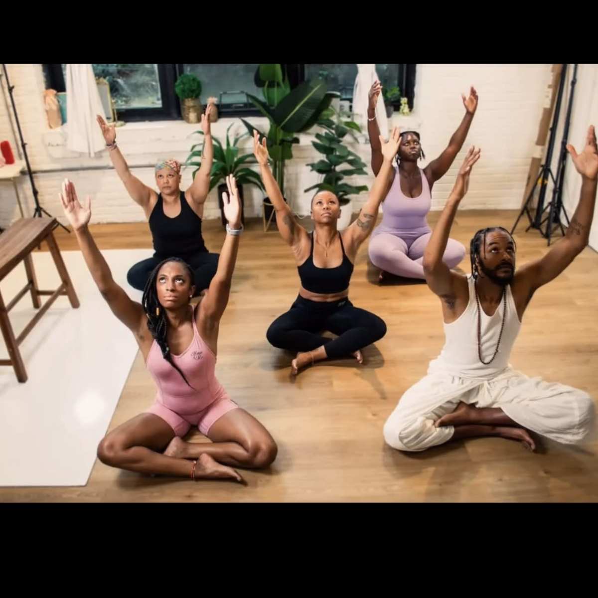 A group of five individuals is practicing yoga in a studio with their arms raised and seated in a cross-legged position.