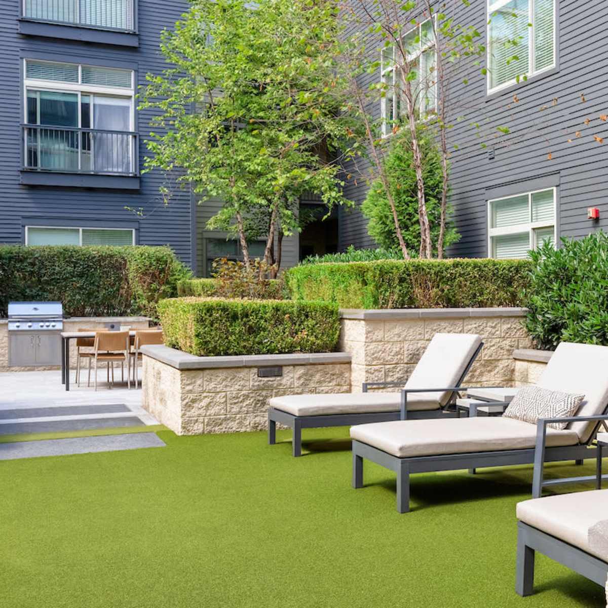 A landscaped courtyard area with lounge chairs, a barbecue grill, and a dining table amidst greenery and stone features.
