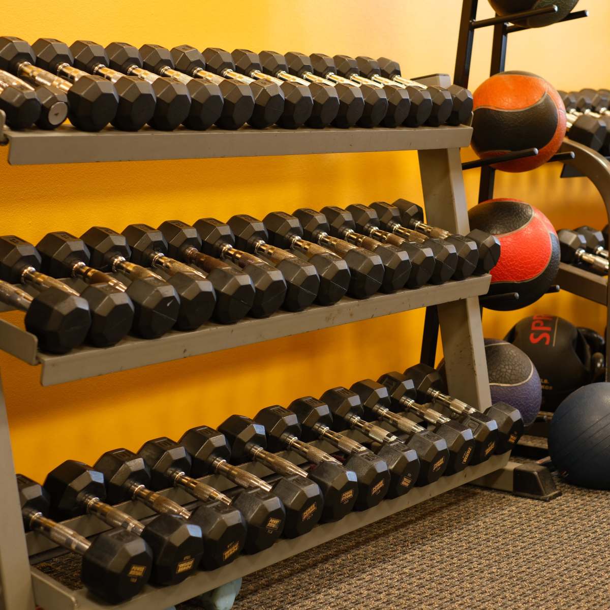 A gym storage area with organized rows of dumbbells, kettlebells, and medicine balls against a yellow wall.