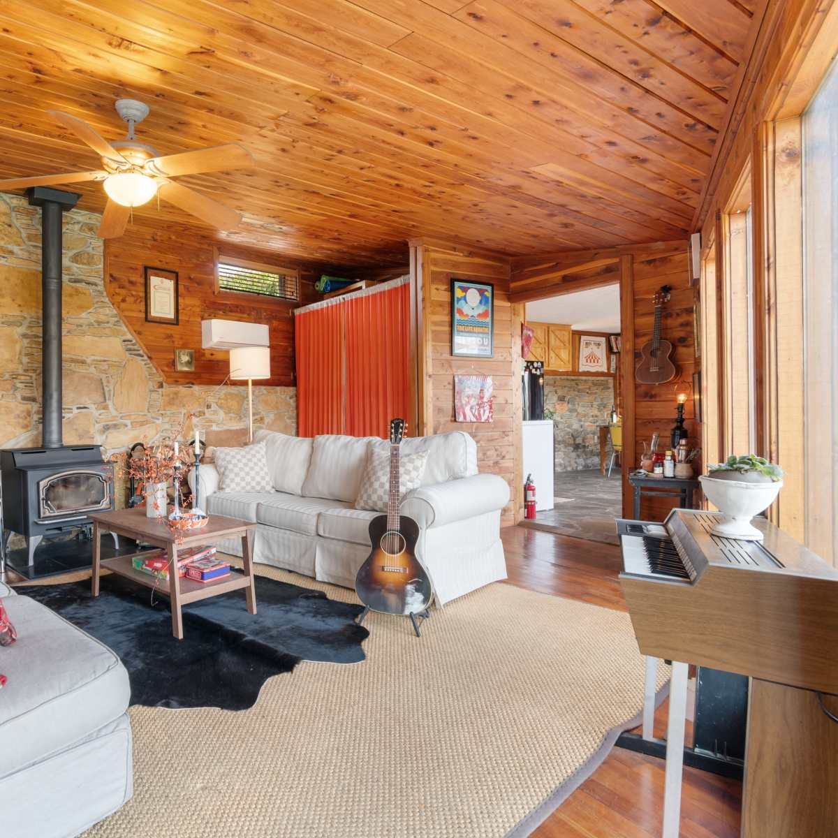 A cozy living room with wooden walls and ceiling, featuring a stone fireplace, two light-colored sofas, and a guitar resting on a black rug near a coffee table.