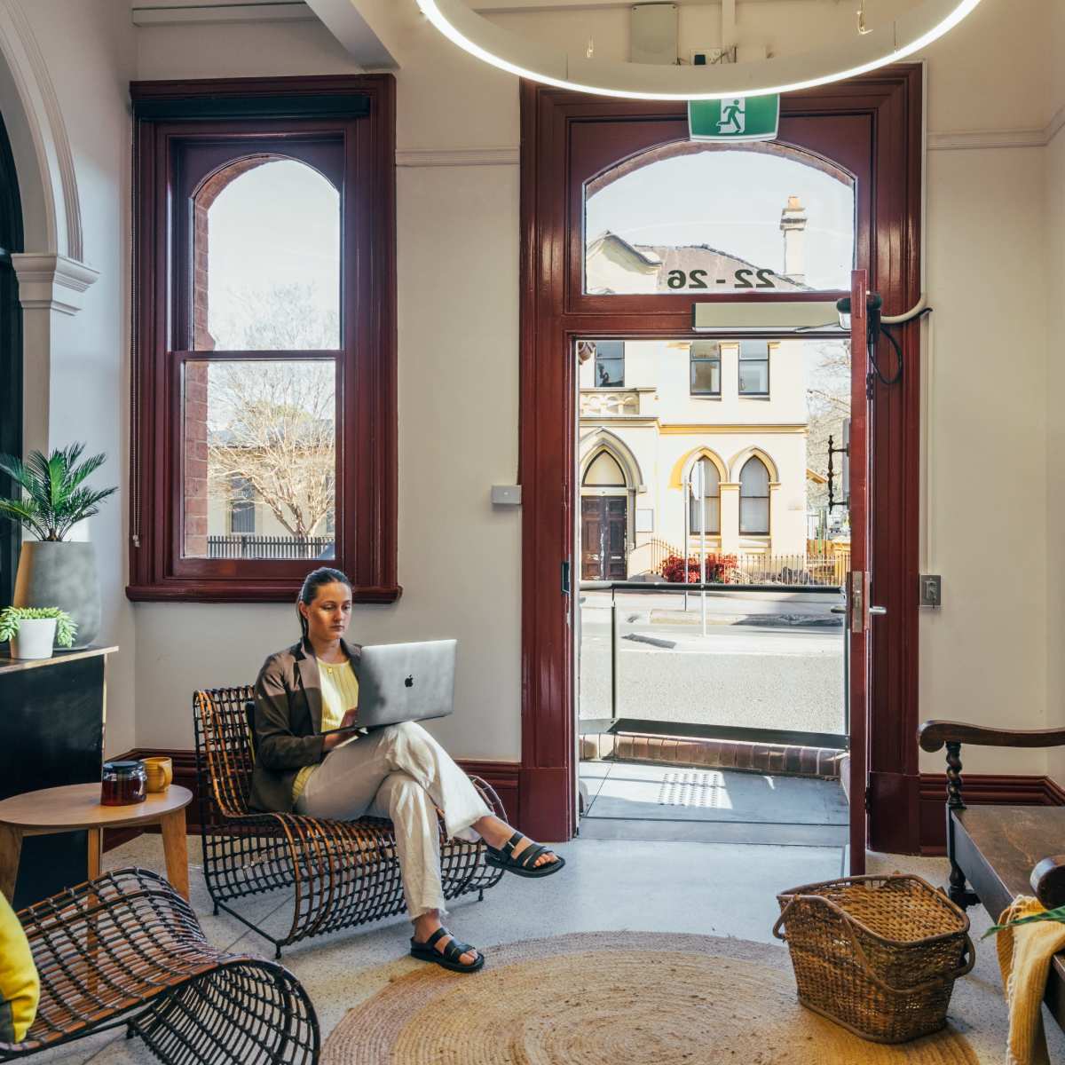 A woman sits on a chair with a laptop in a room featuring large windows and a view of a street outside.