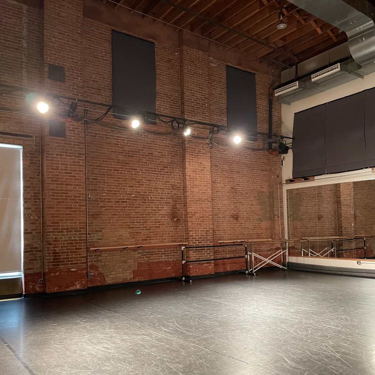 A spacious dance studio with exposed brick walls, overhead lights, and a mirrored wall.