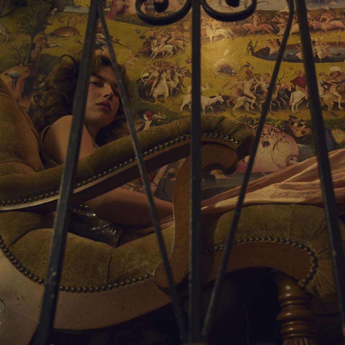 A woman in a low-cut dress sits in a large green armchair in front of a colorful mural depicting various mythical creatures and scenes.