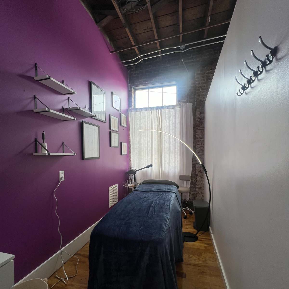 A treatment room with a purple wall, a massage table covered with a dark blanket, and shelves displaying framed pictures and decor.