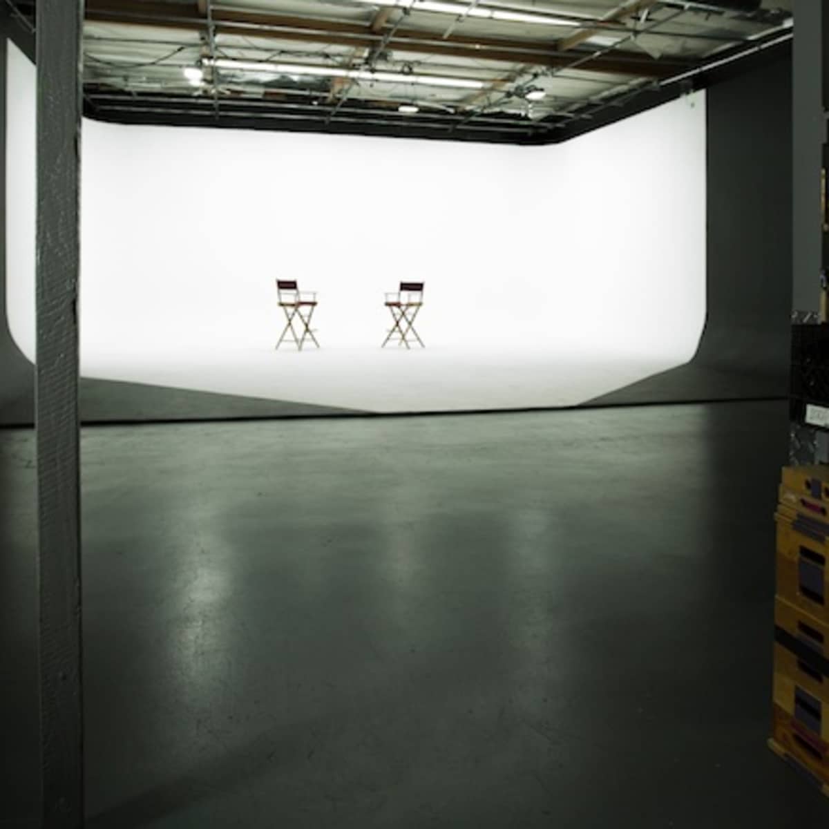 An empty studio space with two director's chairs facing a white backdrop, flanked by shelves containing equipment.