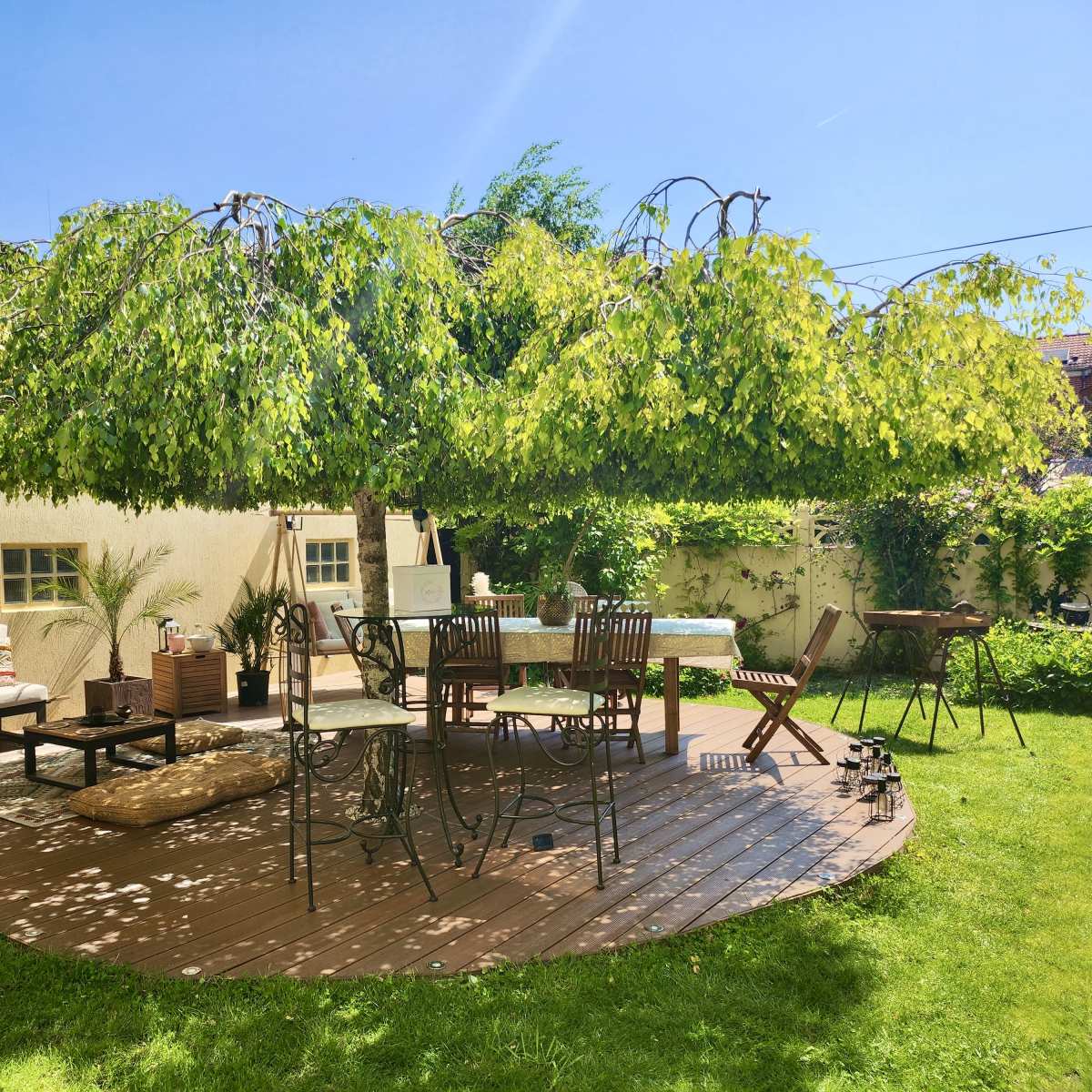A wooden deck features outdoor furniture under a wide canopy of greenery in a well-maintained backyard.