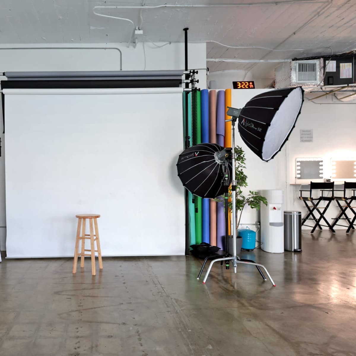 A photography studio with a white backdrop, a wooden stool, and lighting equipment on a polished concrete floor.
