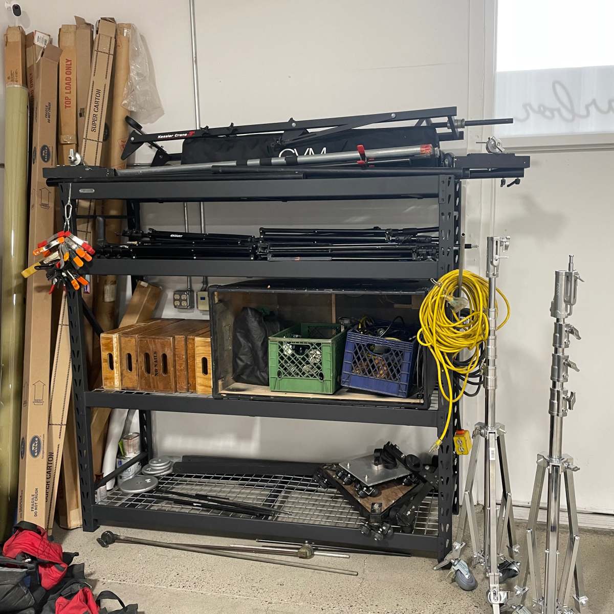 A metal shelving unit in a garage holds various equipment, including tripods, crates, and rolls of cables, alongside a white door with a window.