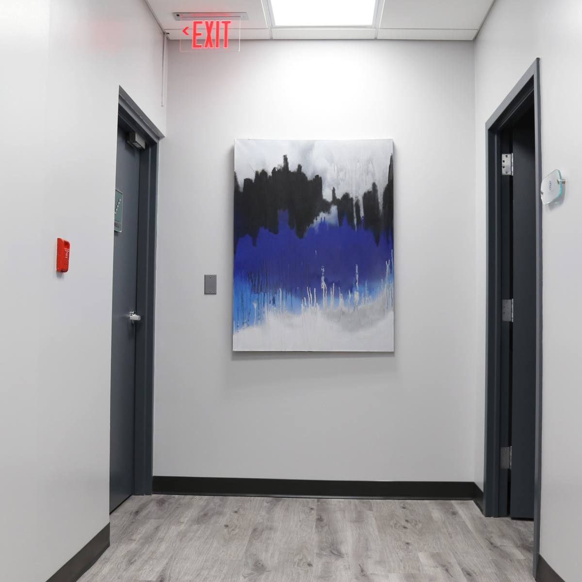 A hallway with grey walls, a wooden floor, and an abstract painting featuring blue, black, and white colors mounted on the wall between two doors.