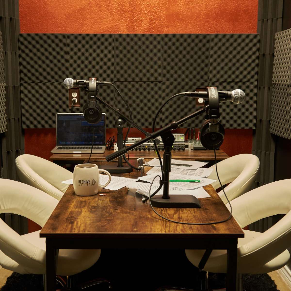 A small recording studio with two microphones, headphones, a laptop, and a table surrounded by acoustic foam panels.