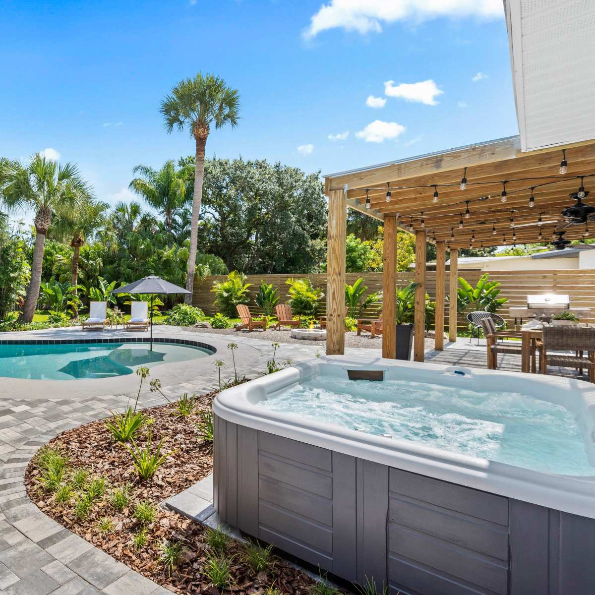 A landscaped backyard with a swimming pool, a hot tub, and a covered seating area surrounded by palm trees and greenery.